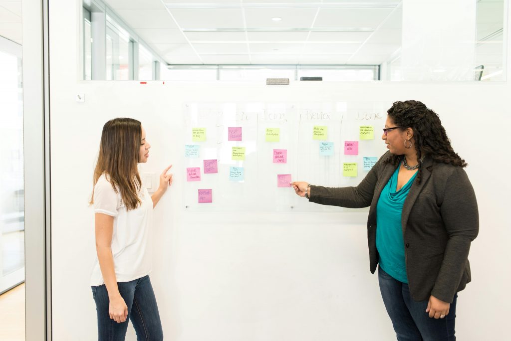 Two women discussing in an office with sticky notes on a whiteboard, showcasing teamwork.