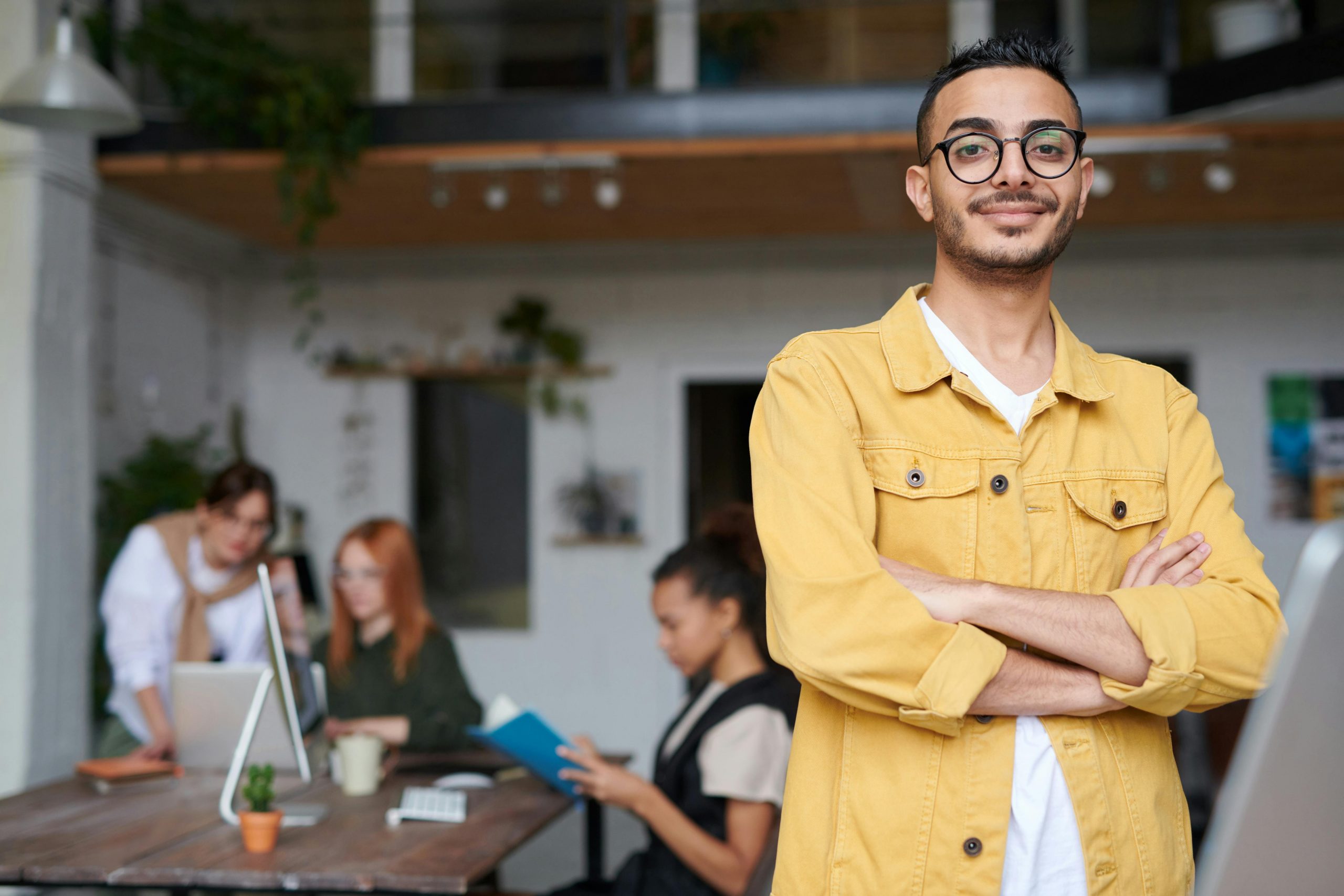 Confident young man in a yellow jacket posing in a modern office environment with colleagues.
